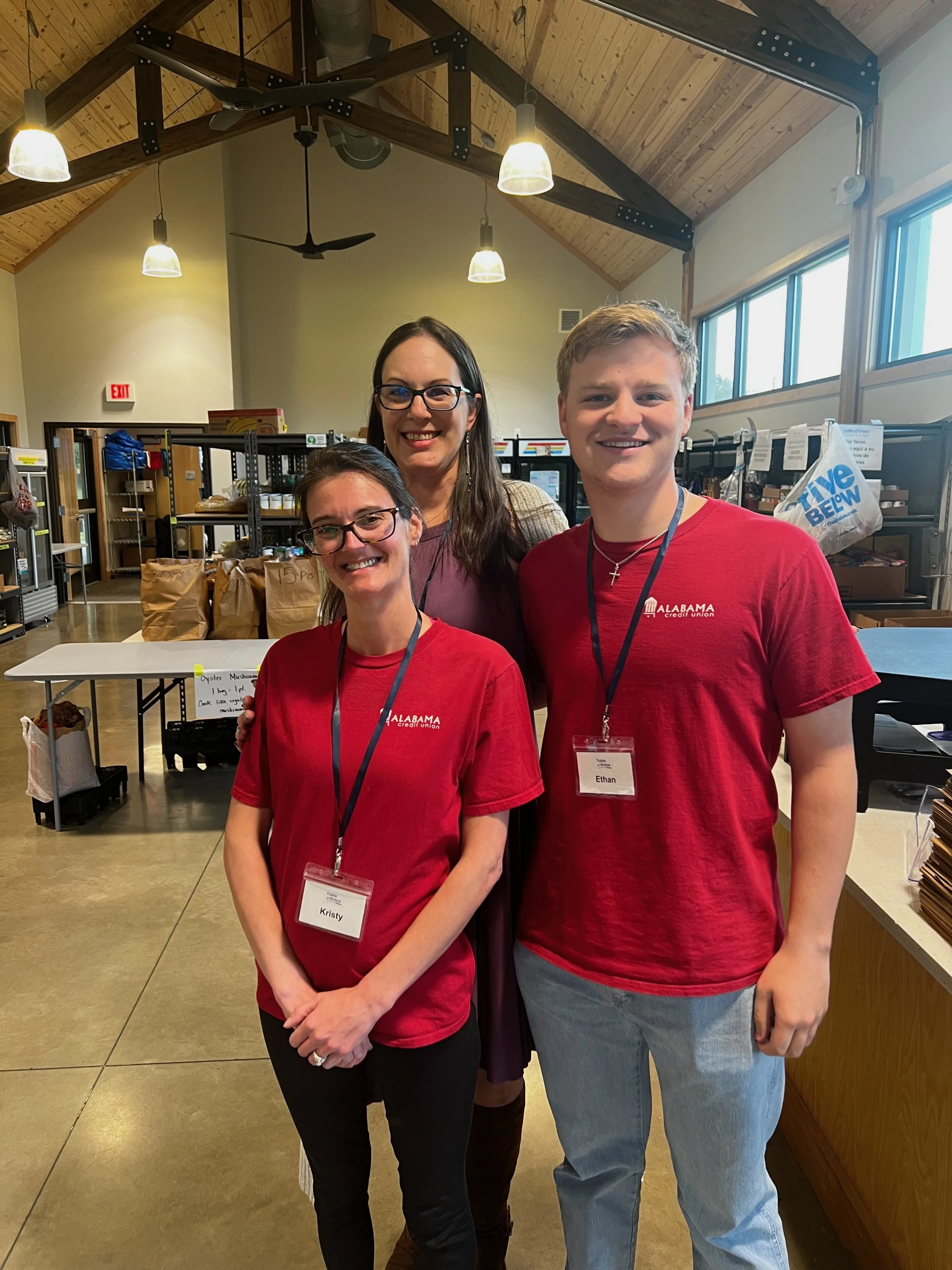 Three people smiling for a photo inside a room with shelves and bags in the background, two wearing red Alabama credit union T-shirts with name tags, and a woman standing behind them.