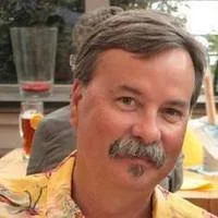 A man with short dark hair, a mustache, and a goatee; smiling at the camera at an outdoor setting with drinks and tables.
