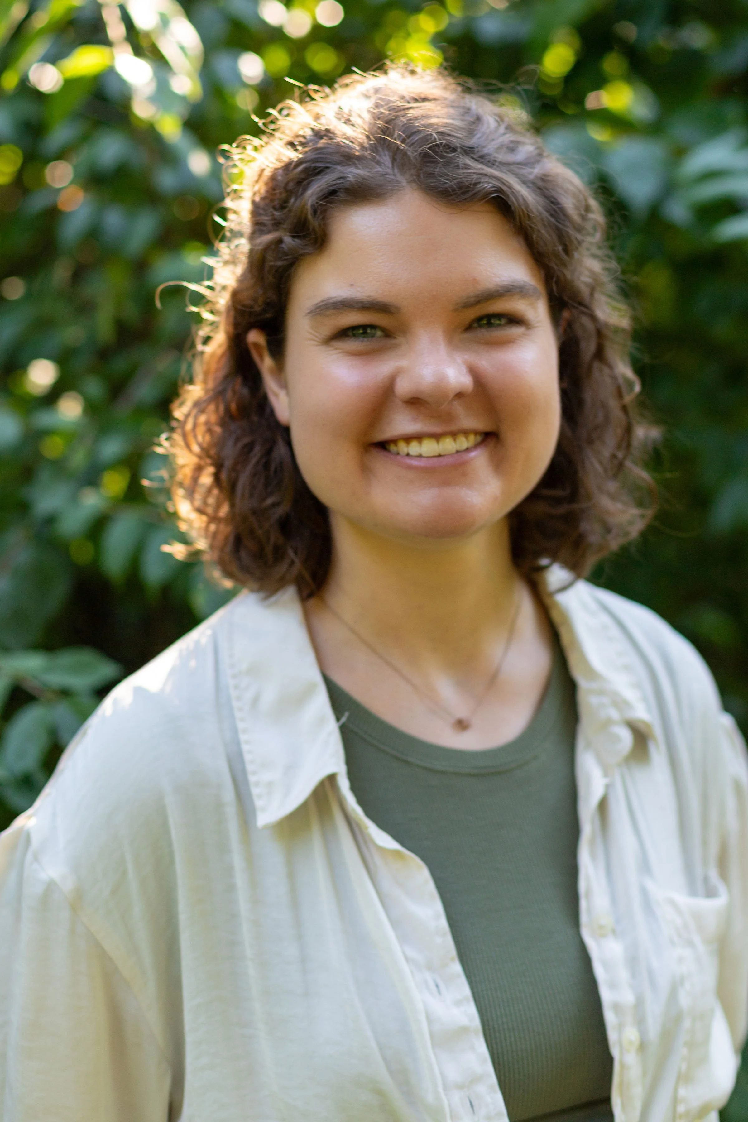 A young woman with curly brown hair smiling outdoors with lush green foliage in the background.