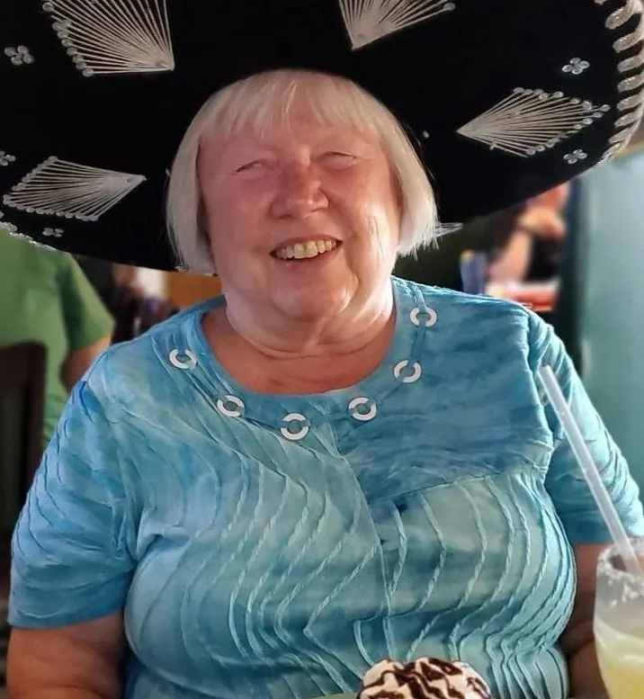 An elderly woman smiling, wearing a large black sombrero decorated with white designs, a light blue shirt with a textured pattern, and a necklace. She has a dessert with whipped cream and chocolate drizzle in front of her.