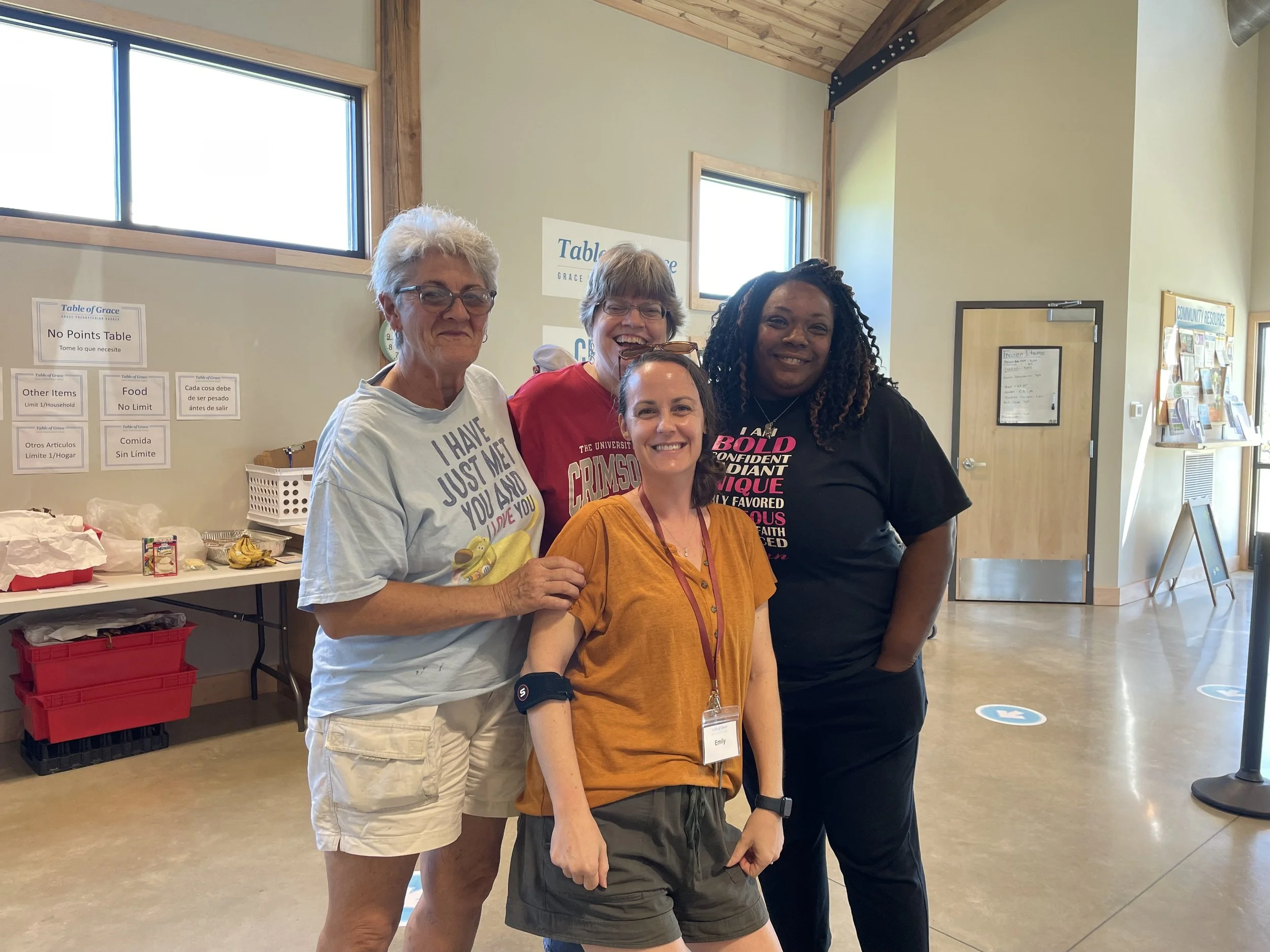 Four women standing together in a well-lit indoor space, smiling at the camera. Behind them is a table with various items, and posters and signs on the walls.