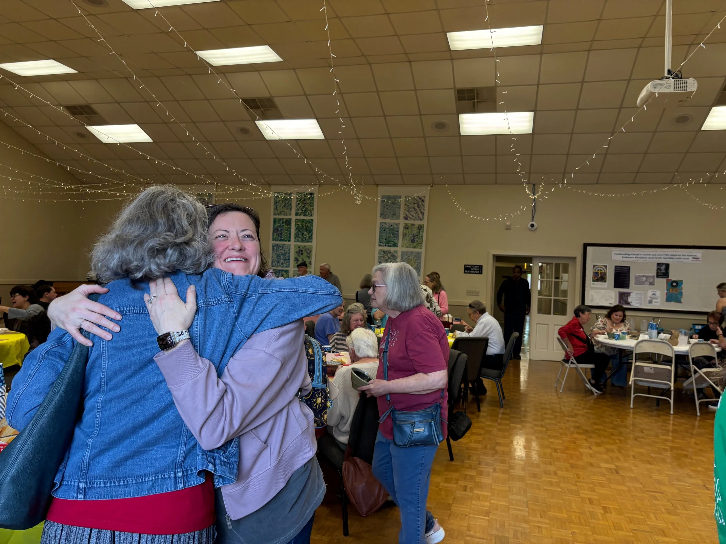 Two women hugging and smiling in a gathering hall filled with people sitting at tables, autumn decorations, and string lights hanging from the ceiling.