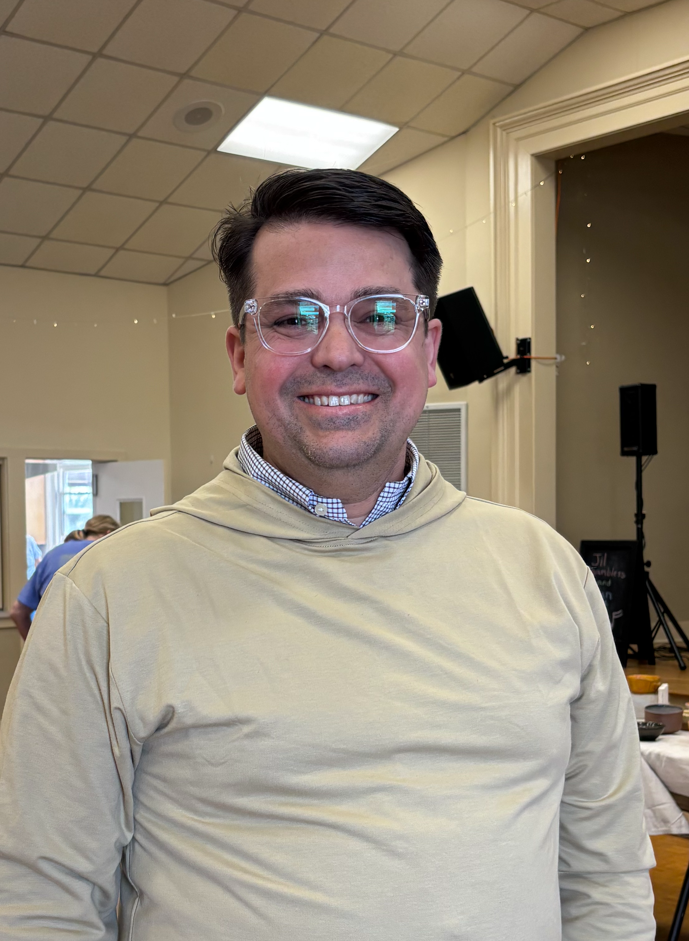 Smiling man with glasses and a beige hoodie inside a room with a ceiling light and some black speakers.
