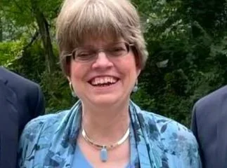 A young man sitting on a beige cushioned bench against a plain wall, wearing a gray polo shirt and cream-colored pants, with a serious expression and a shadow cast on the wall.