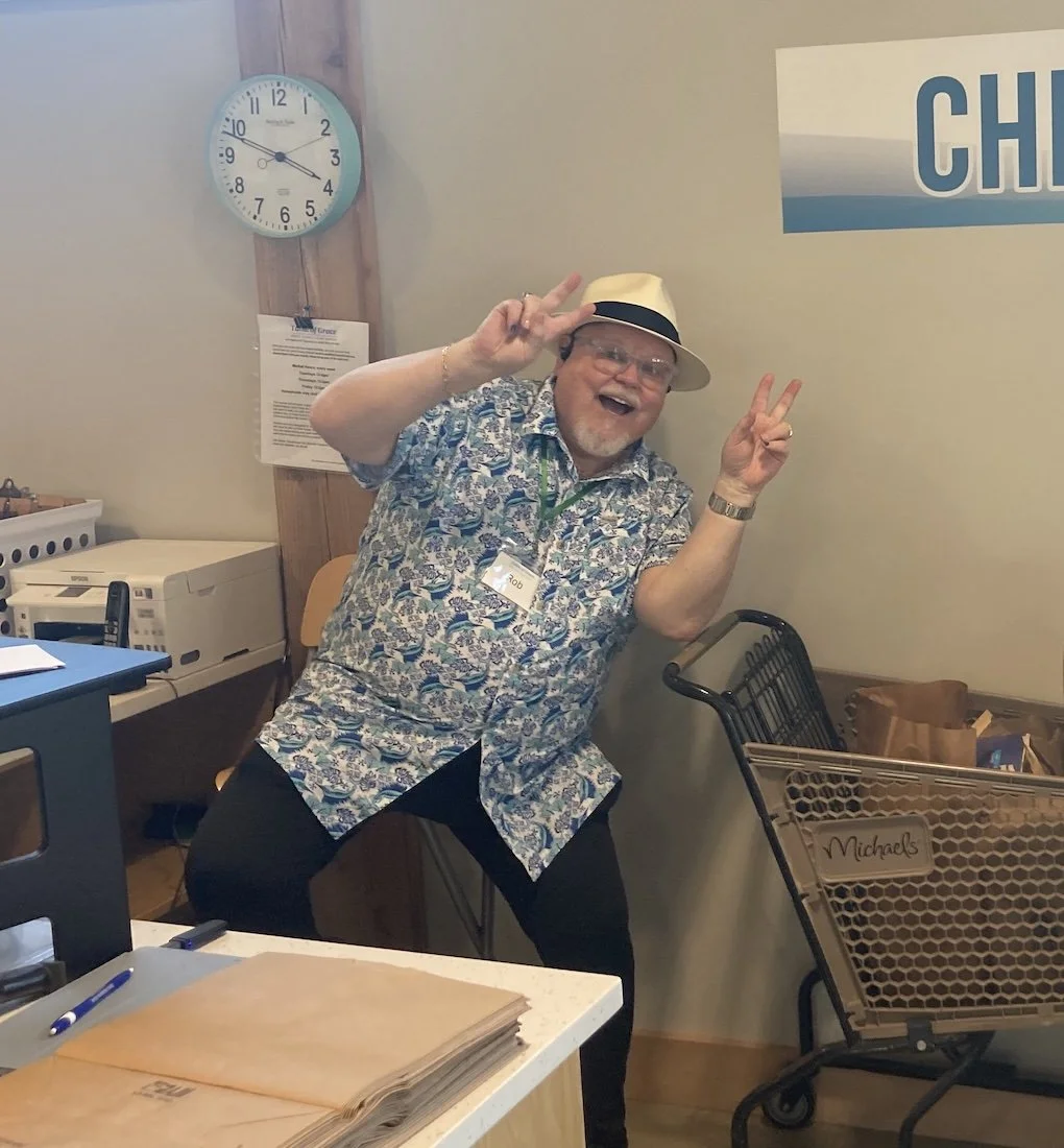 A cheerful man with glasses, a hat, and a patterned shirt making peace signs near a wall with a clock and a poster at an office or store.