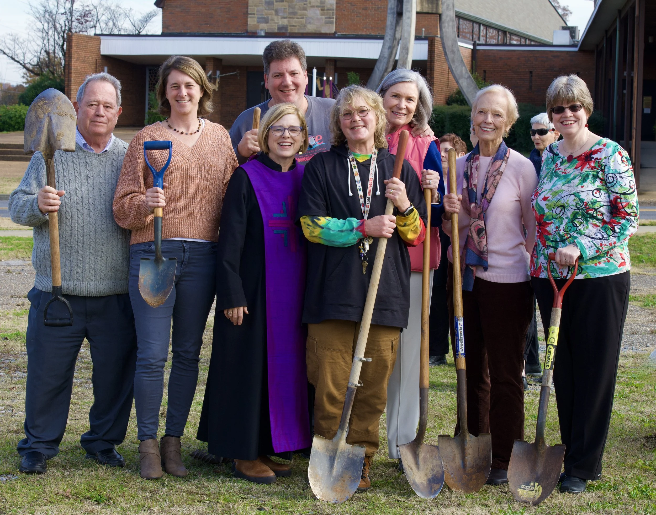 Group of ten diverse adults outdoors holding gardening tools, smiling, standing on grass in front of a building with a tree.