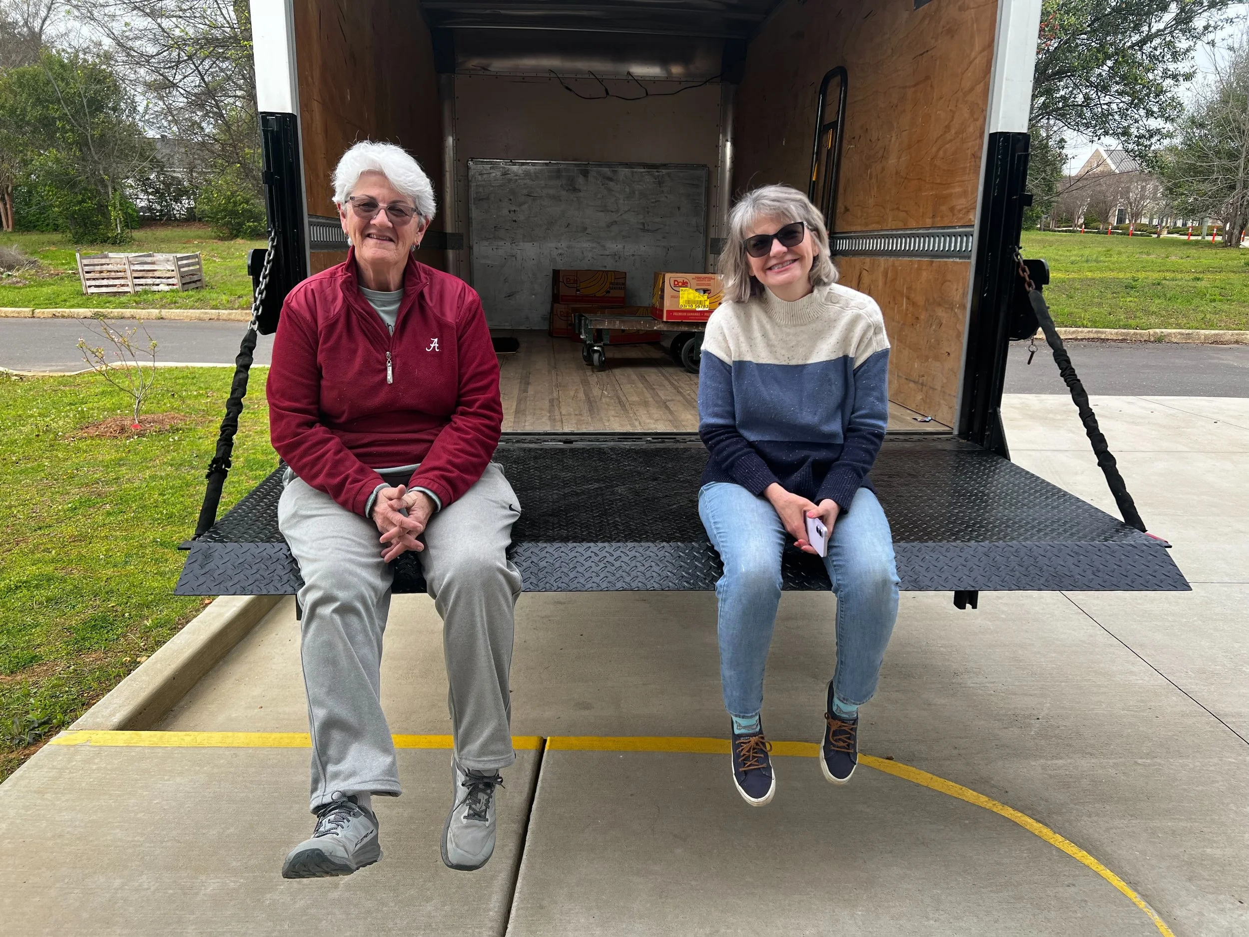 Two women sitting on a swing attached to a moving truck. The woman on the left has white hair, glasses, and is wearing a red jacket and gray pants. The woman on the right has gray hair, sunglasses, a color-blocked sweater, and jeans. They are outdoors in a parking lot with grass and trees in the background.