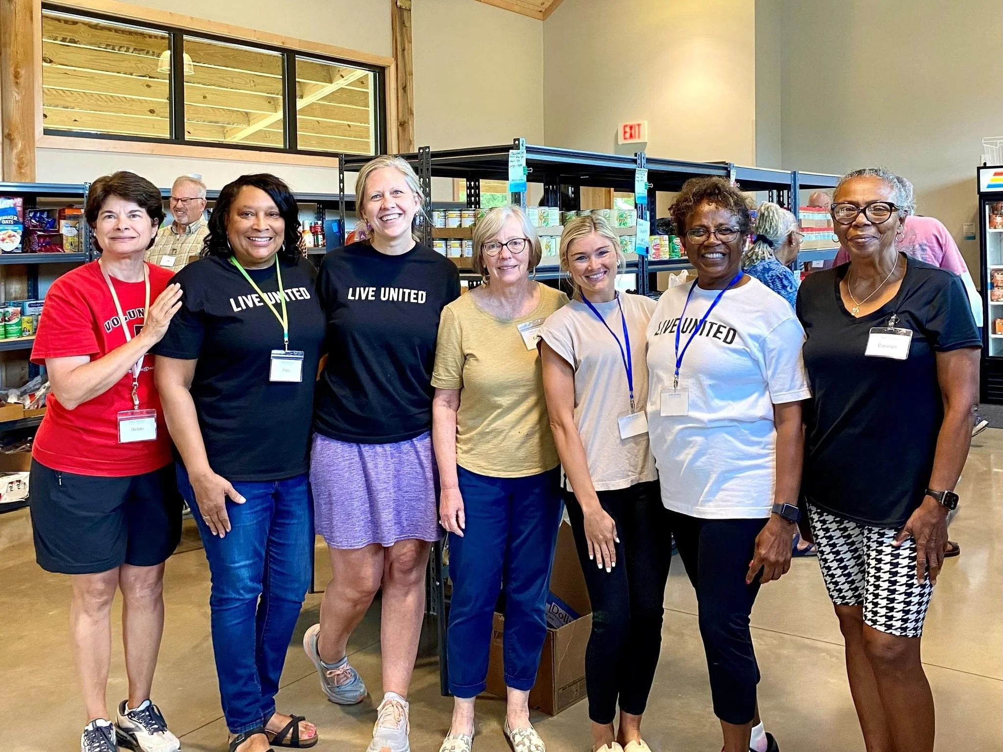 Group of seven women standing inside a store aisle, some wearing 'LIVE UNITED' shirts, smiling at the camera.