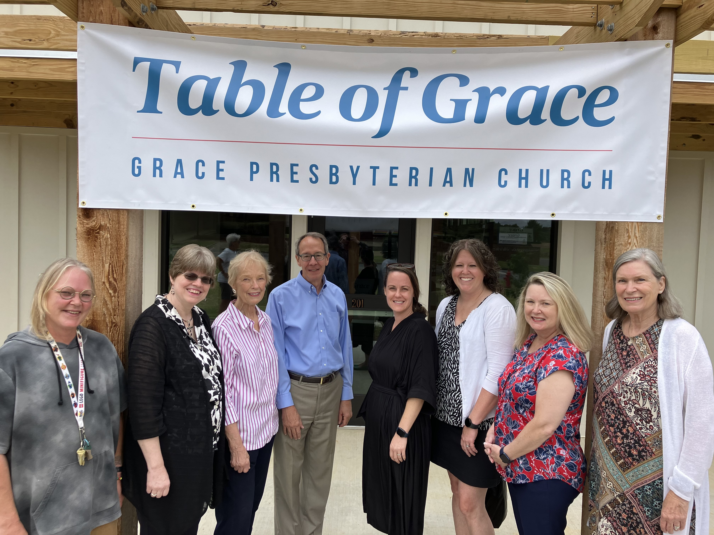 Group of nine diverse adults standing in front of a church building under a large banner that reads 'Table of Grace, Grace Presbyterian Church'.