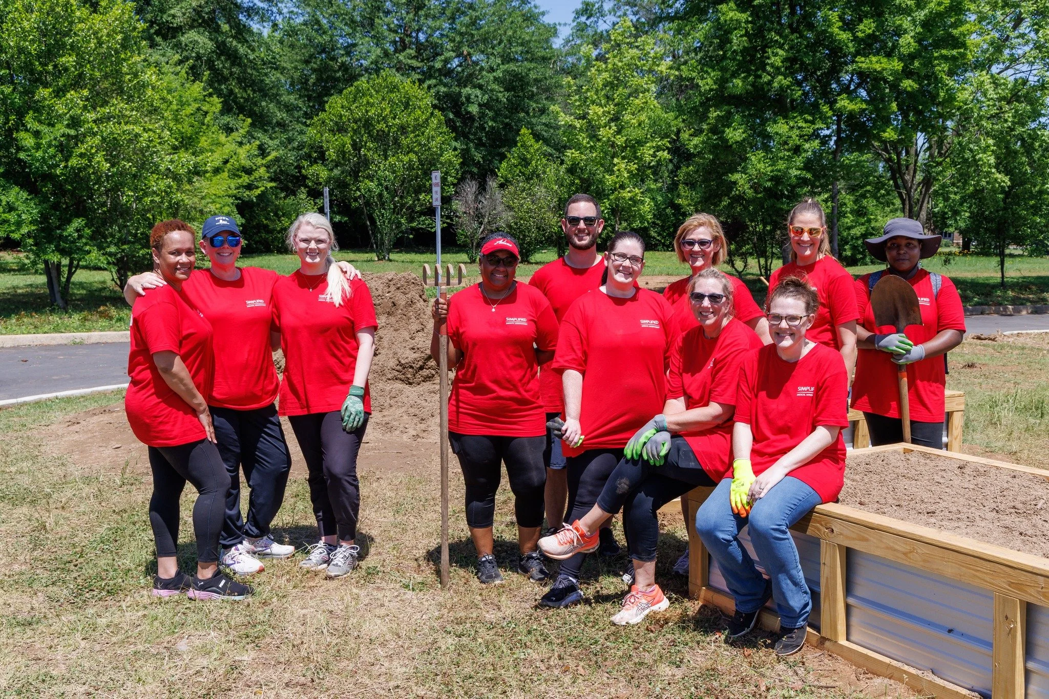 Group of volunteers in red shirts posing outdoors with a raised garden bed and a shovel during a community planting event.