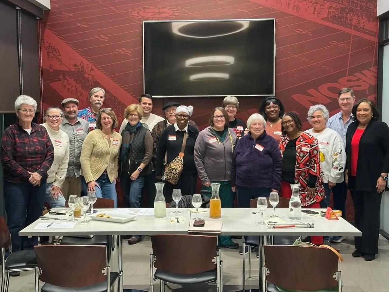 Group of 16 diverse people standing together in a dining room, smiling at the camera, with a large TV screen on a red wall behind them, and a table with drinks and glasses in front.