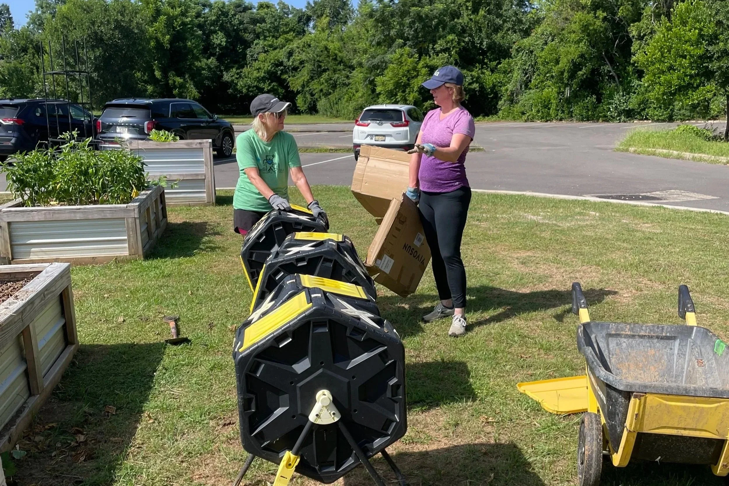 Two women working in a garden planting vegetables, with one woman holding a cardboard box and the other woman standing nearby.
