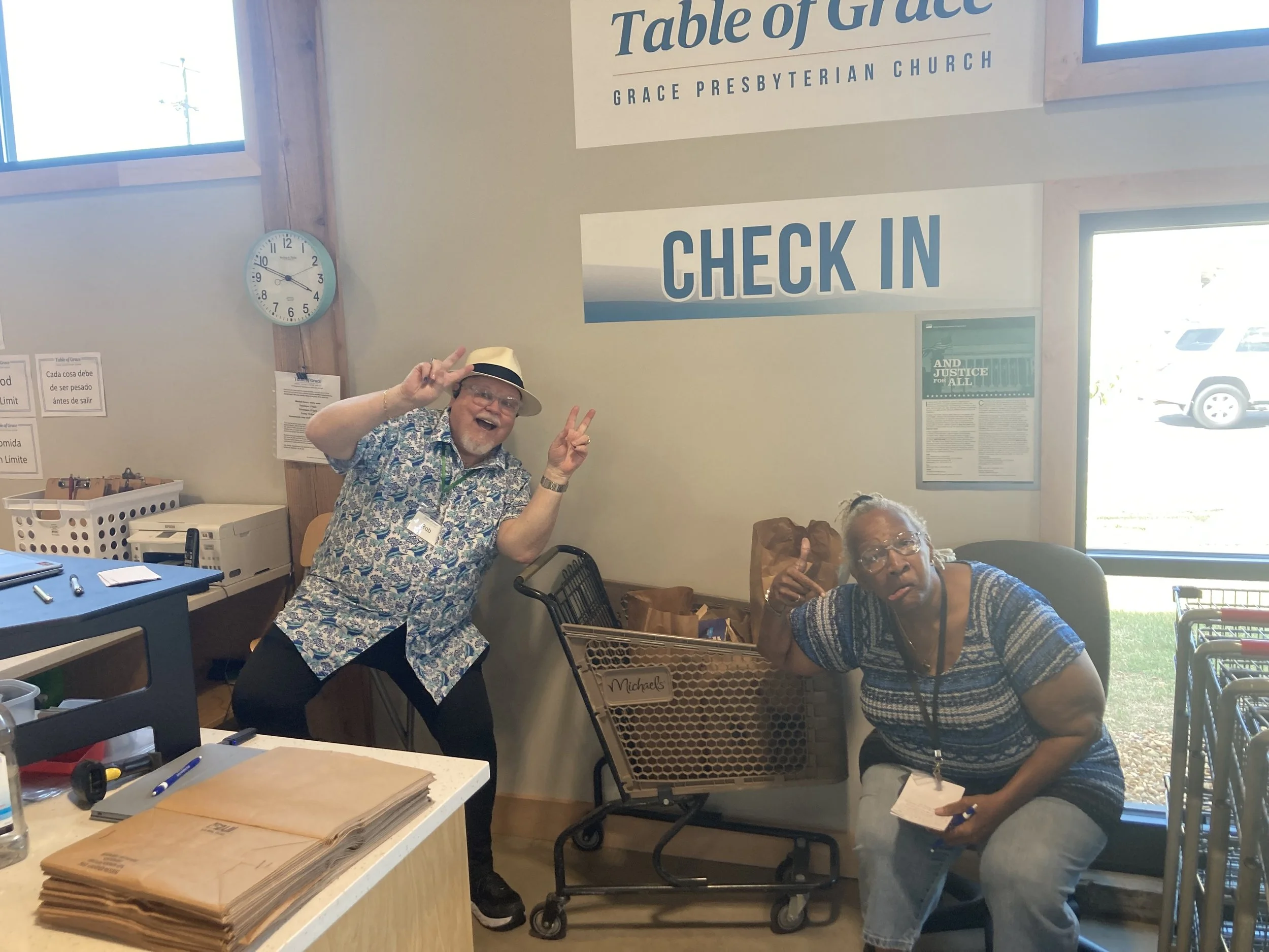 Two smiling older individuals making peace signs at a check-in counter in a church, with a sign that says 'Table of Grace' and 'Check In' behind them, in a well-lit room with a window showing parked cars outside.