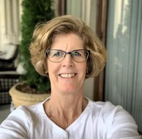 A woman with glasses and short, curly blonde hair smiling at the camera, outdoors near a building with a potted plant in the background.