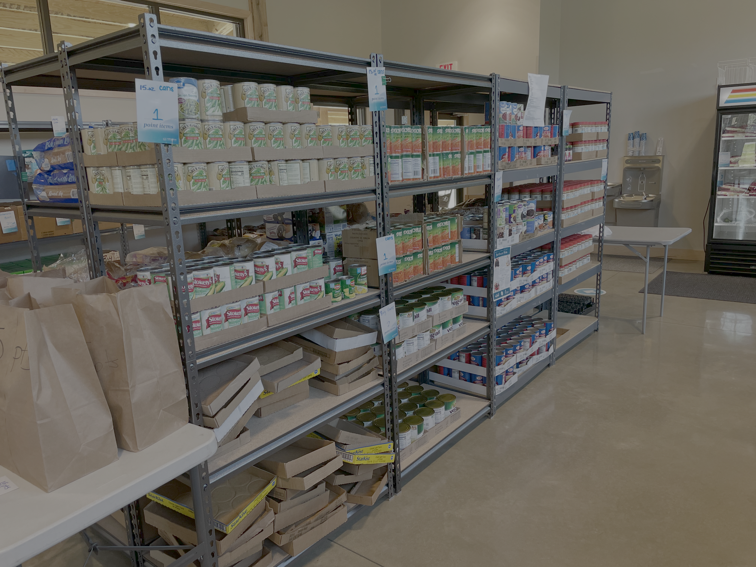 Shelves stocked with canned food in a food pantry or grocery store, with paper bags and boxes on a table nearby.