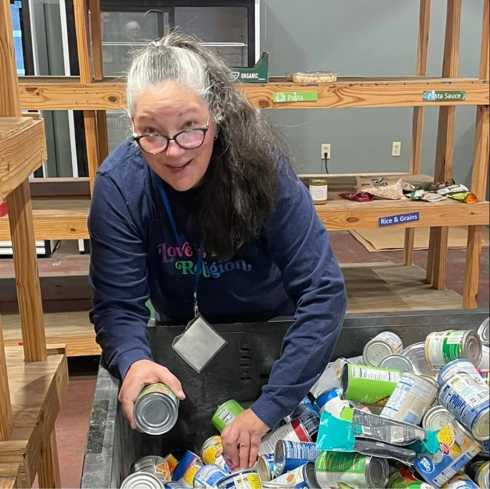 A woman with glasses and gray hair with dark hair, wearing a navy blue shirt, is leaning over a large bin of canned goods, sorting or organizing them. She is in a room with wooden shelves labeled for different food items.