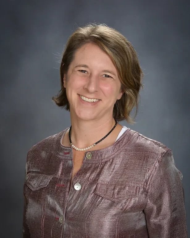 A smiling woman with short light brown hair, wearing a textured gray jacket, a pearl necklace, and a layered black and white shirt underneath, against a dark gray background.