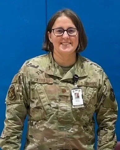Military woman in camouflage uniform standing against a blue wall.