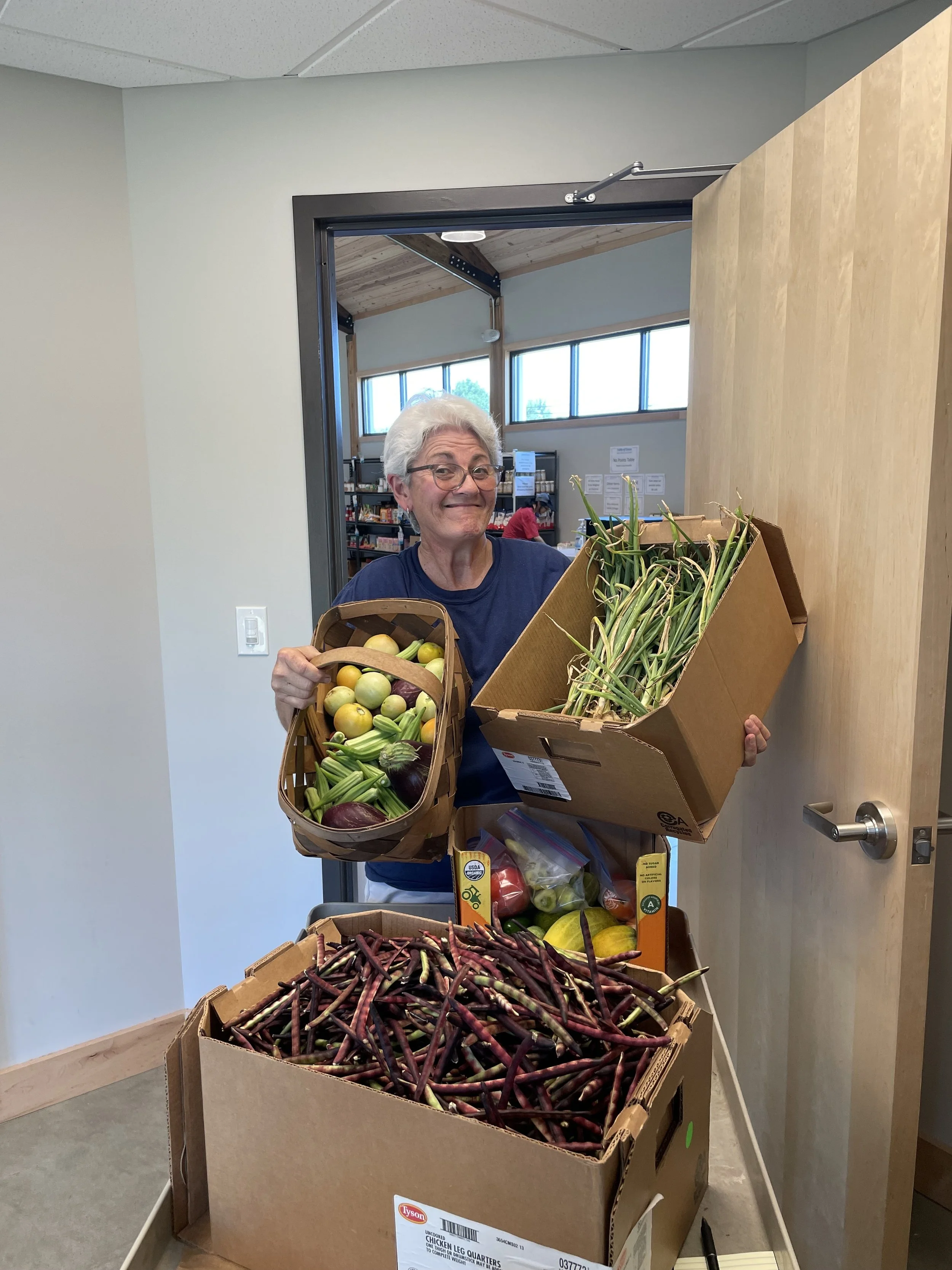 An elderly woman with white hair and glasses smiling while holding baskets and boxes of fresh vegetables inside a room.