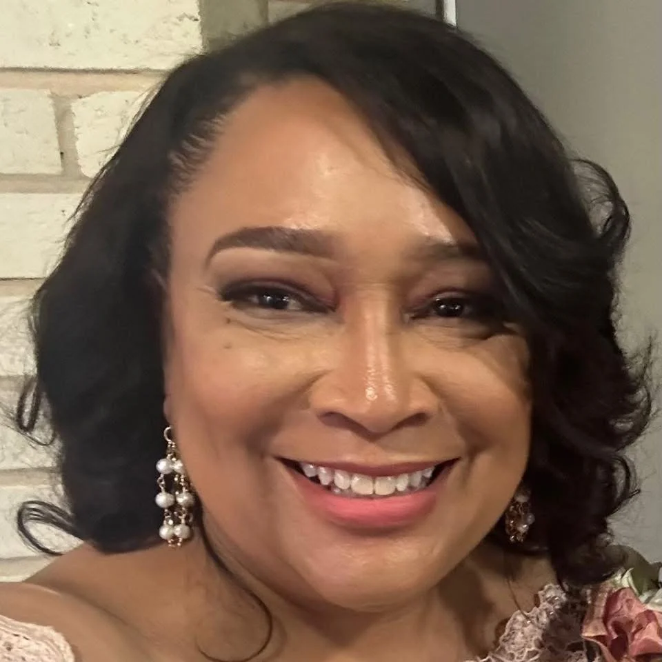 A smiling woman with dark wavy hair, wearing pearl and gold earrings, and makeup, standing in front of a brick wall.