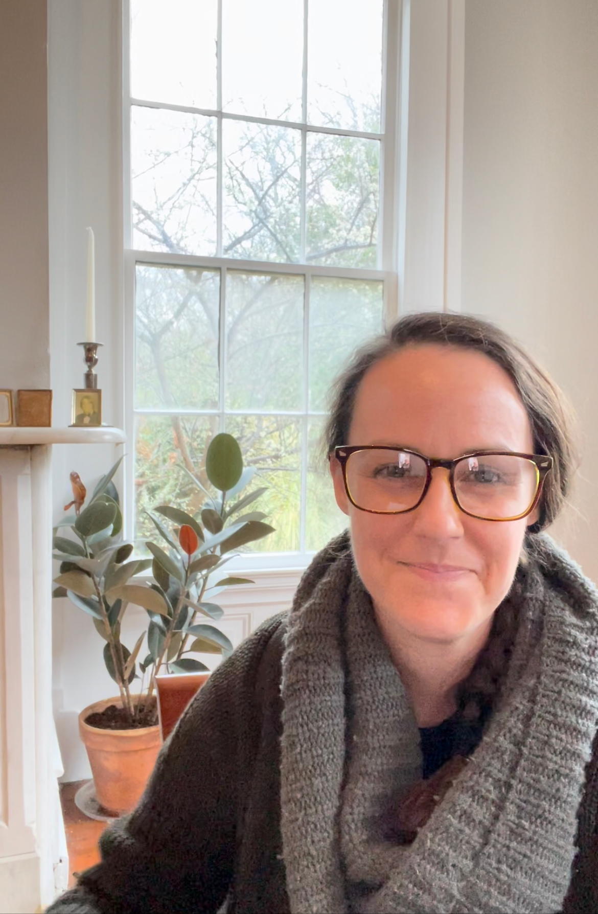 A woman wearing glasses and a grey scarf, smiling, sitting indoors near a window with a plant and small decorative items on the mantelpiece.