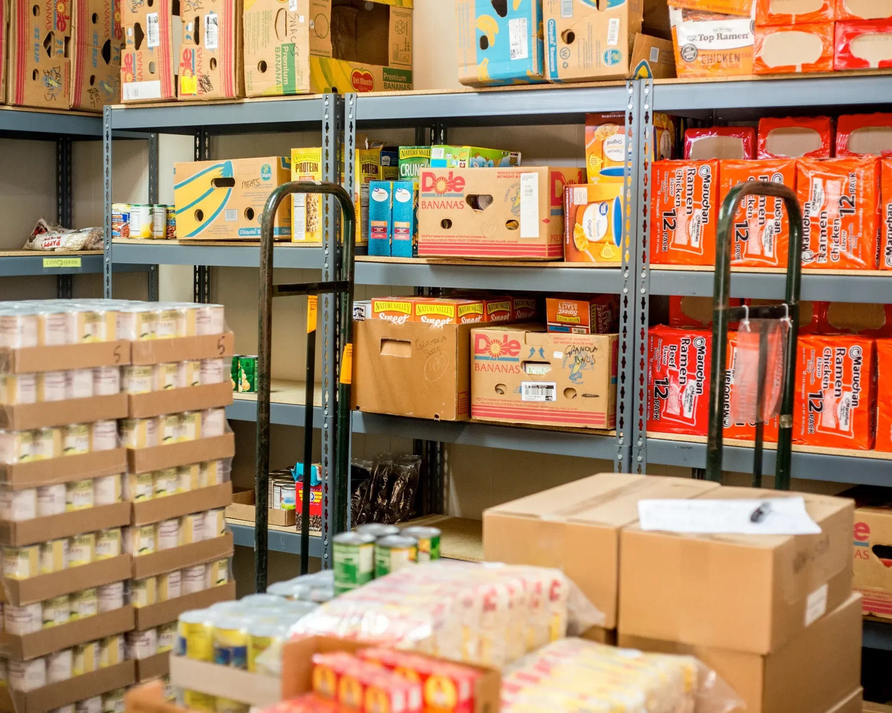 Shelves stocked with boxes of snacks and supplies, including bananas, ramen noodles, canned goods, and packaged items for food storage or distribution in a storage room or pantry.