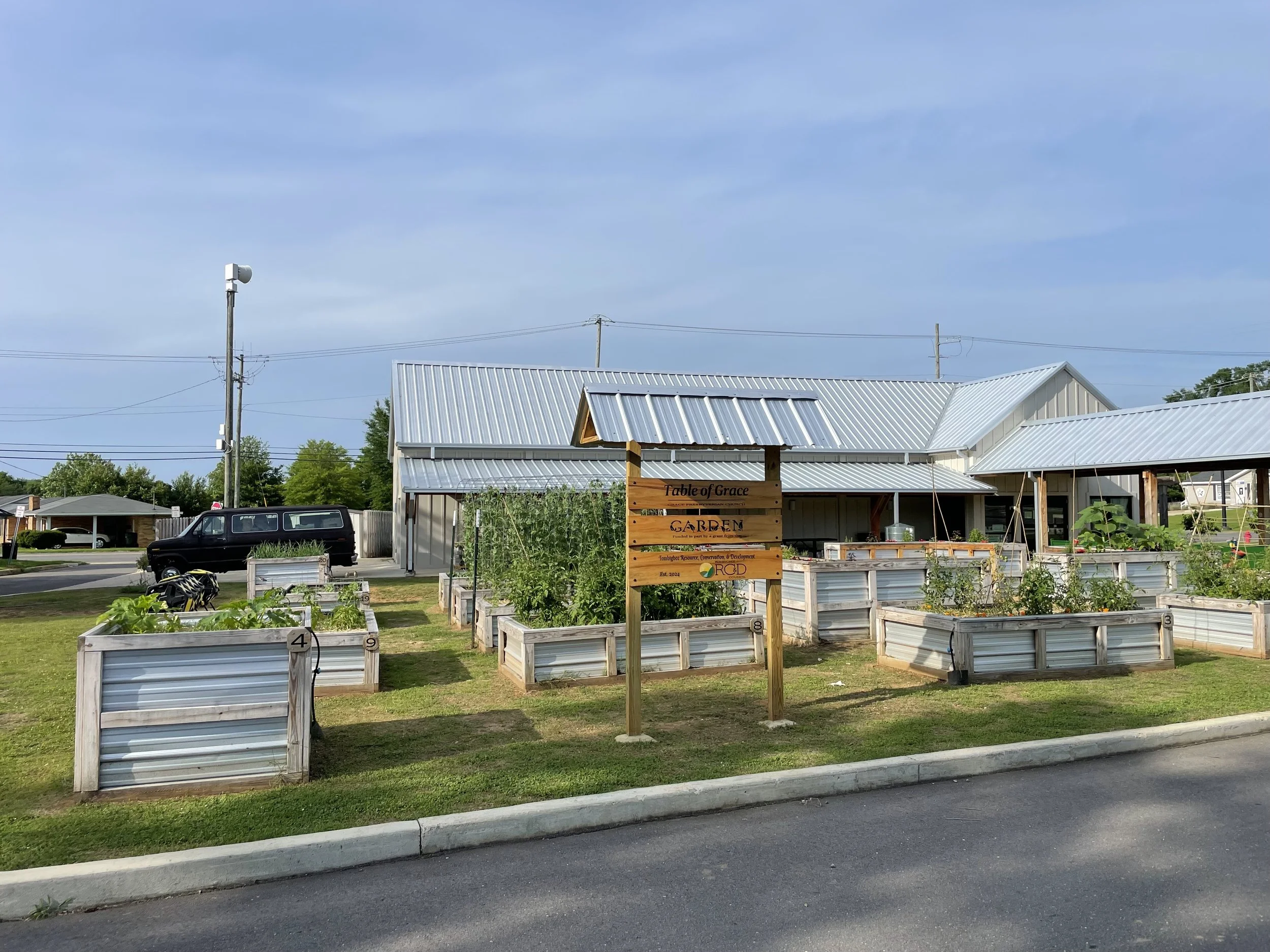 Community garden with raised metal planter boxes, a sign reading 'Table of Grace Garden,' and a building with a metal roof in the background.