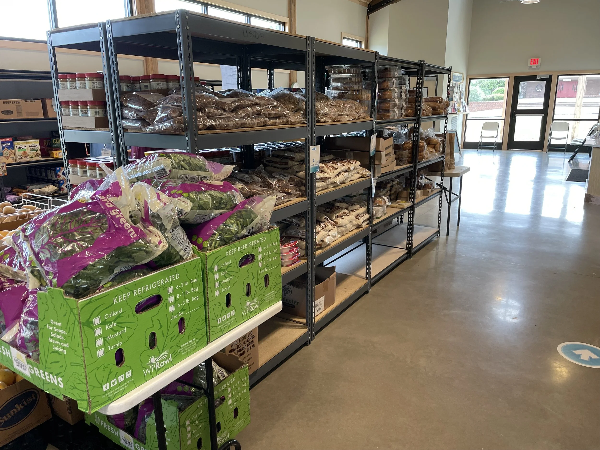 Inside a grocery store with metal shelving units stocked with bags of various grains and produce, near the entrance with glass doors and windows, and a floor marker with an upward arrow.