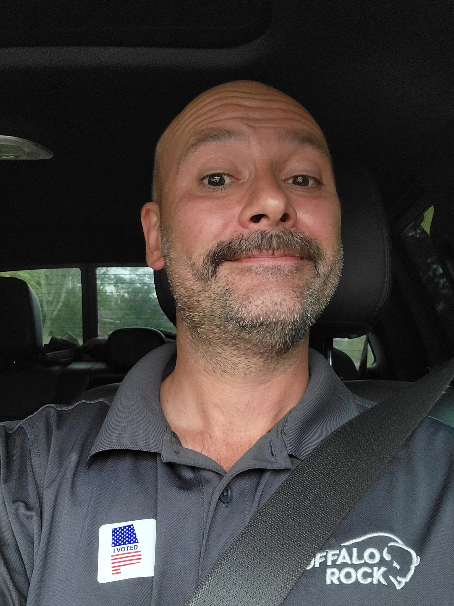 A man with a beard and mustache taking a selfie inside a vehicle, wearing a gray collared shirt with a 'BUFFALO ROCK' logo and an 'I VOTED' sticker.