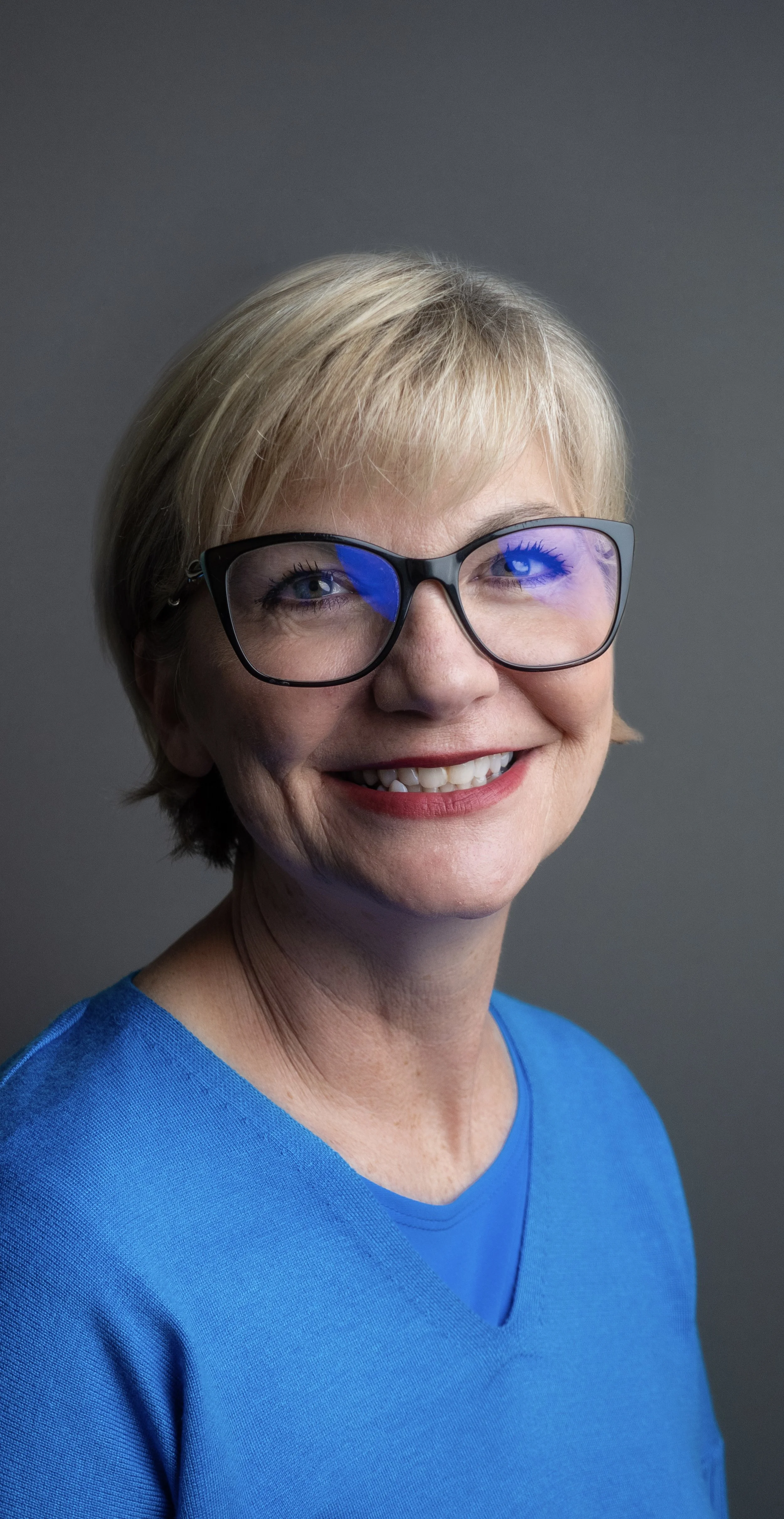 Portrait of a smiling middle-aged woman with short blonde hair, wearing glasses with a reflection of blue light, and a blue shirt against a gray background.