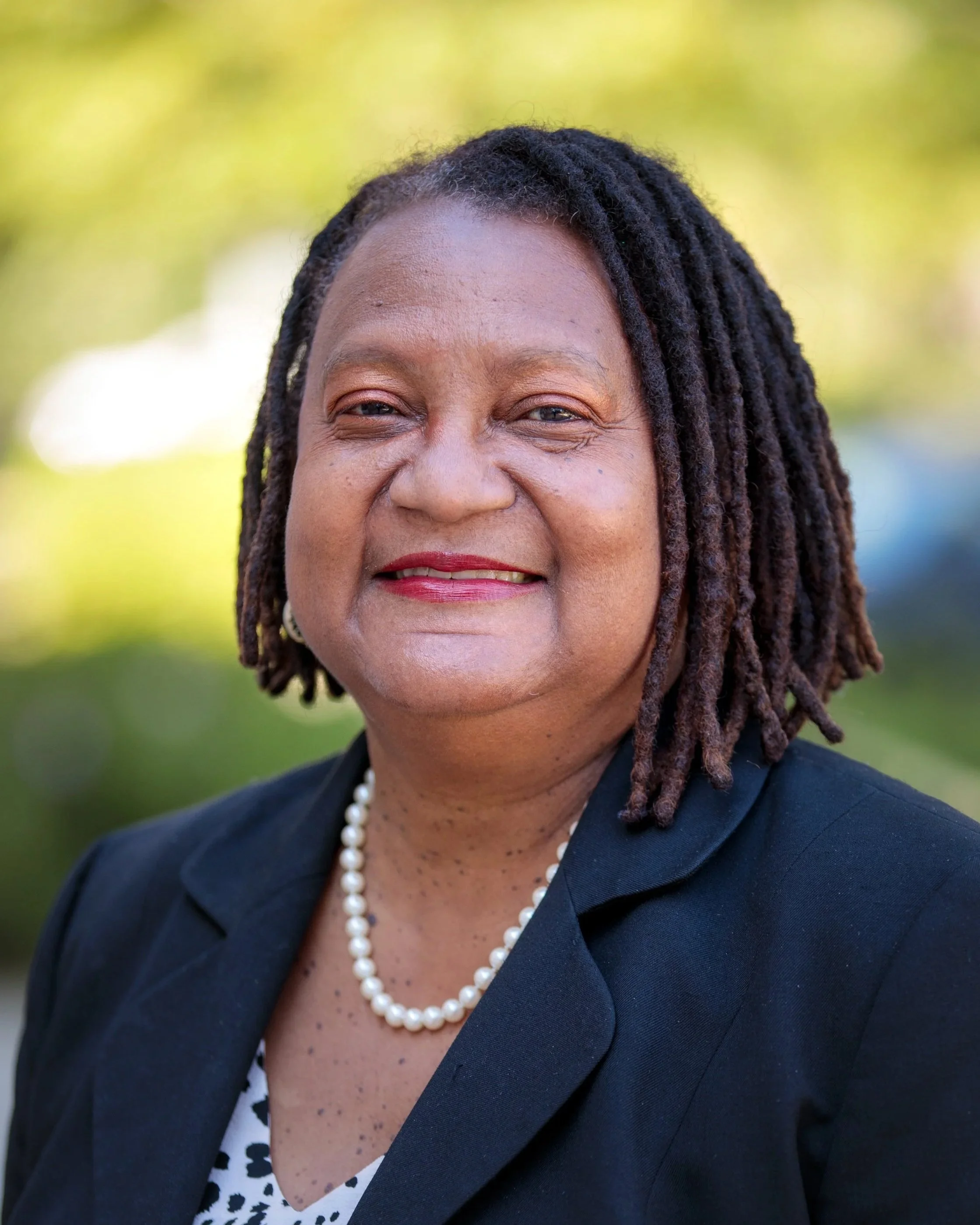 A middle-aged man with a gray beard sitting on a wooden bench against a plain wall, looking contemplative.
