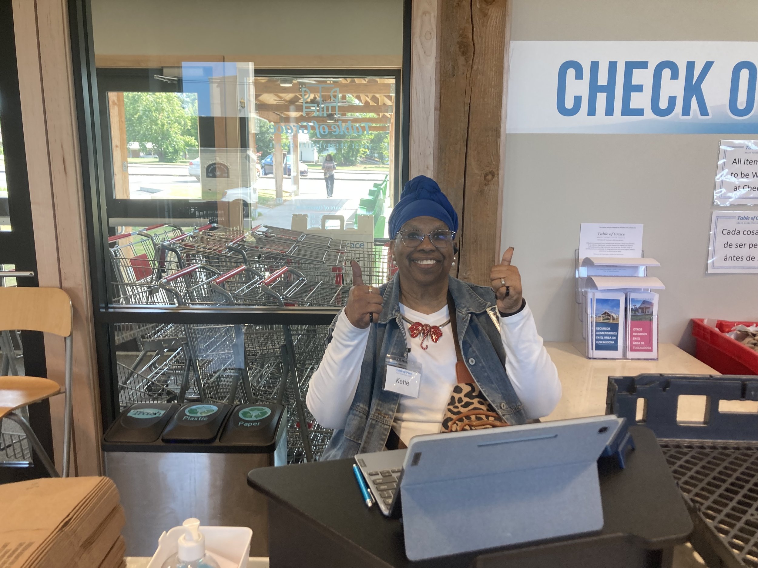 Smiling woman with glasses and blue head wrap giving two thumbs up at checkout counter in grocery store.