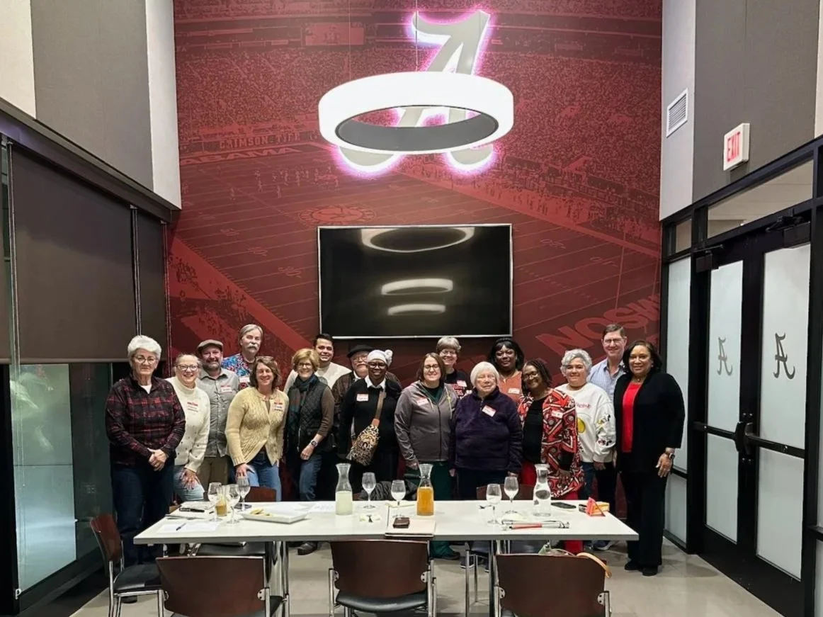 Group of people standing in a restaurant or conference room with a football theme, large flat-screen TV, and a distinctive ring-shaped light fixture hanging from the ceiling. Dining table in front with glasses, pitchers, and plates.