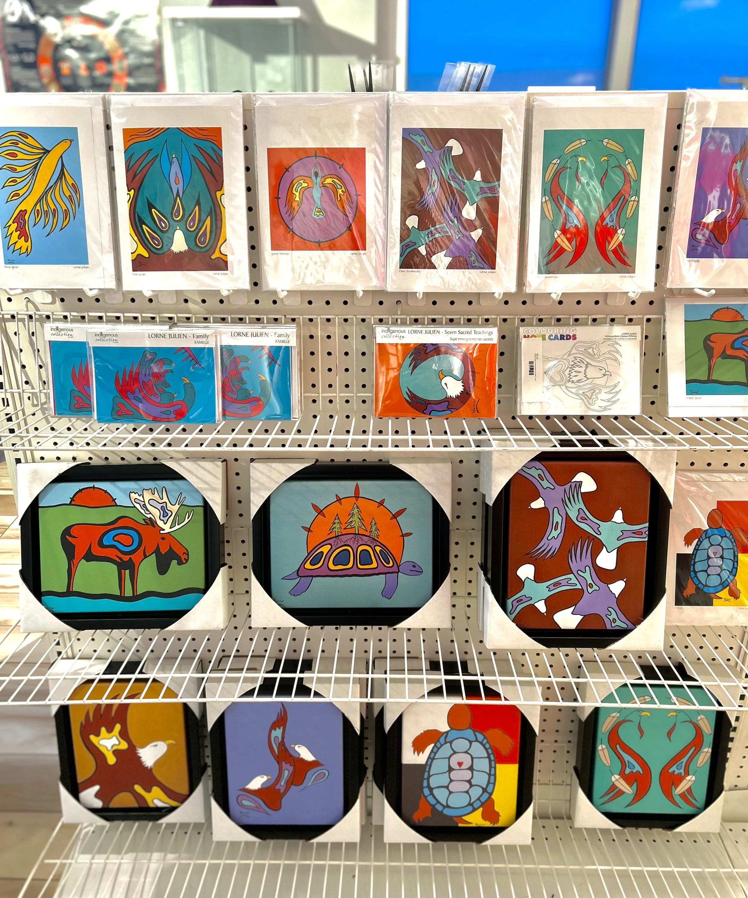 Display of Native American-inspired artwork featuring animals such as turtles, deer, and birds, with vibrant colors and abstract designs arranged on a white wire shelf.
