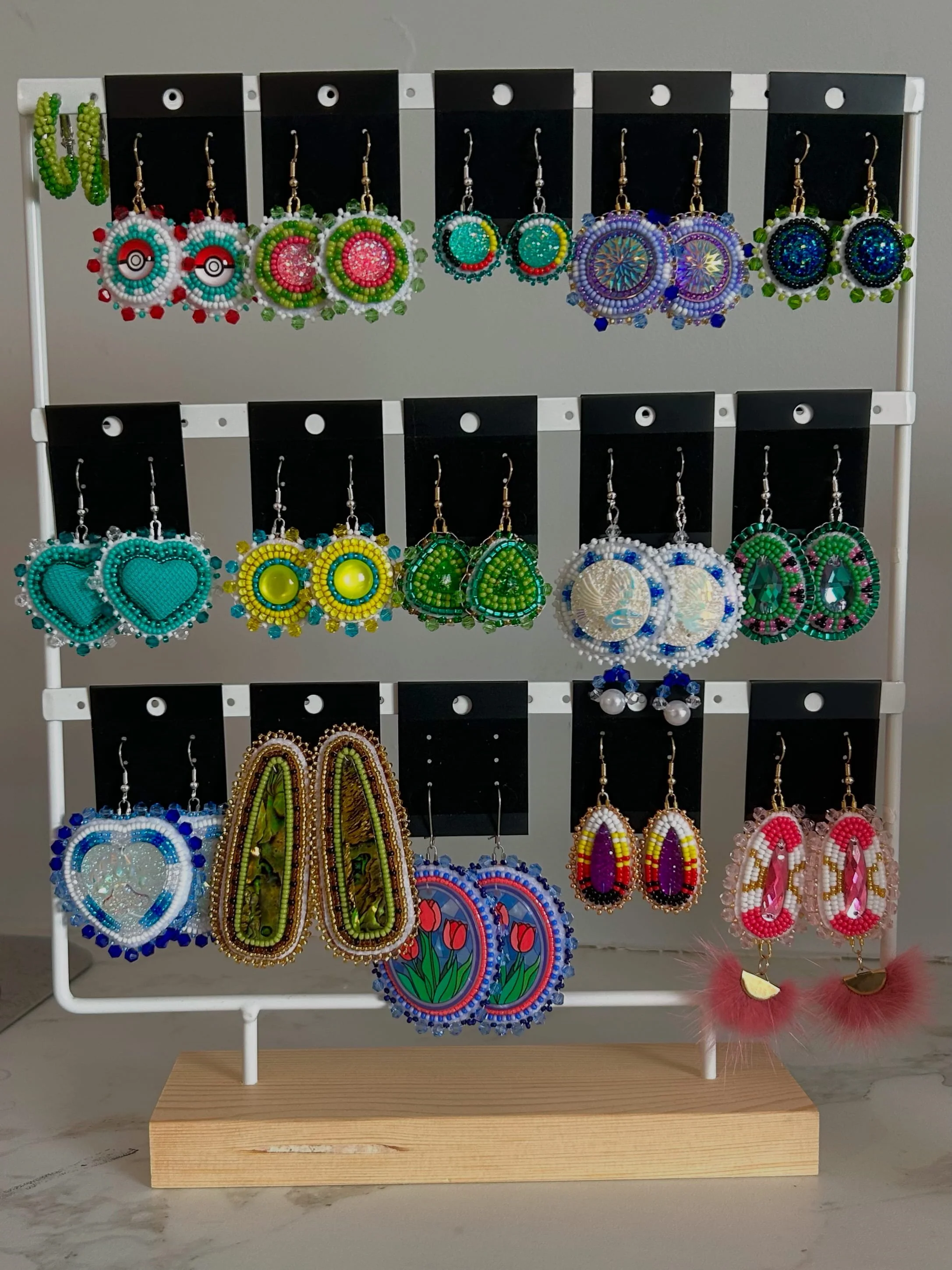 Display stand with colorful beaded and painted earrings arranged on black cards, featuring various shapes like circles, hearts, teardrops, and abstract designs.