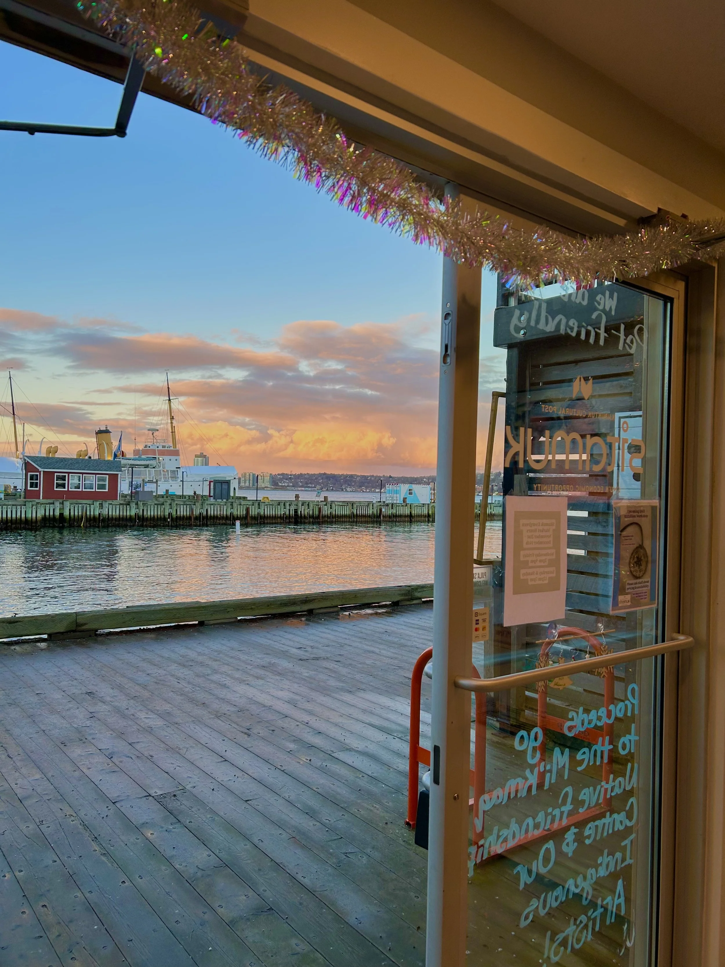 View of a harbor through a doorway, with boats docked and a sunset sky with clouds in the background. The door has a sign and some handwritten text on the glass.