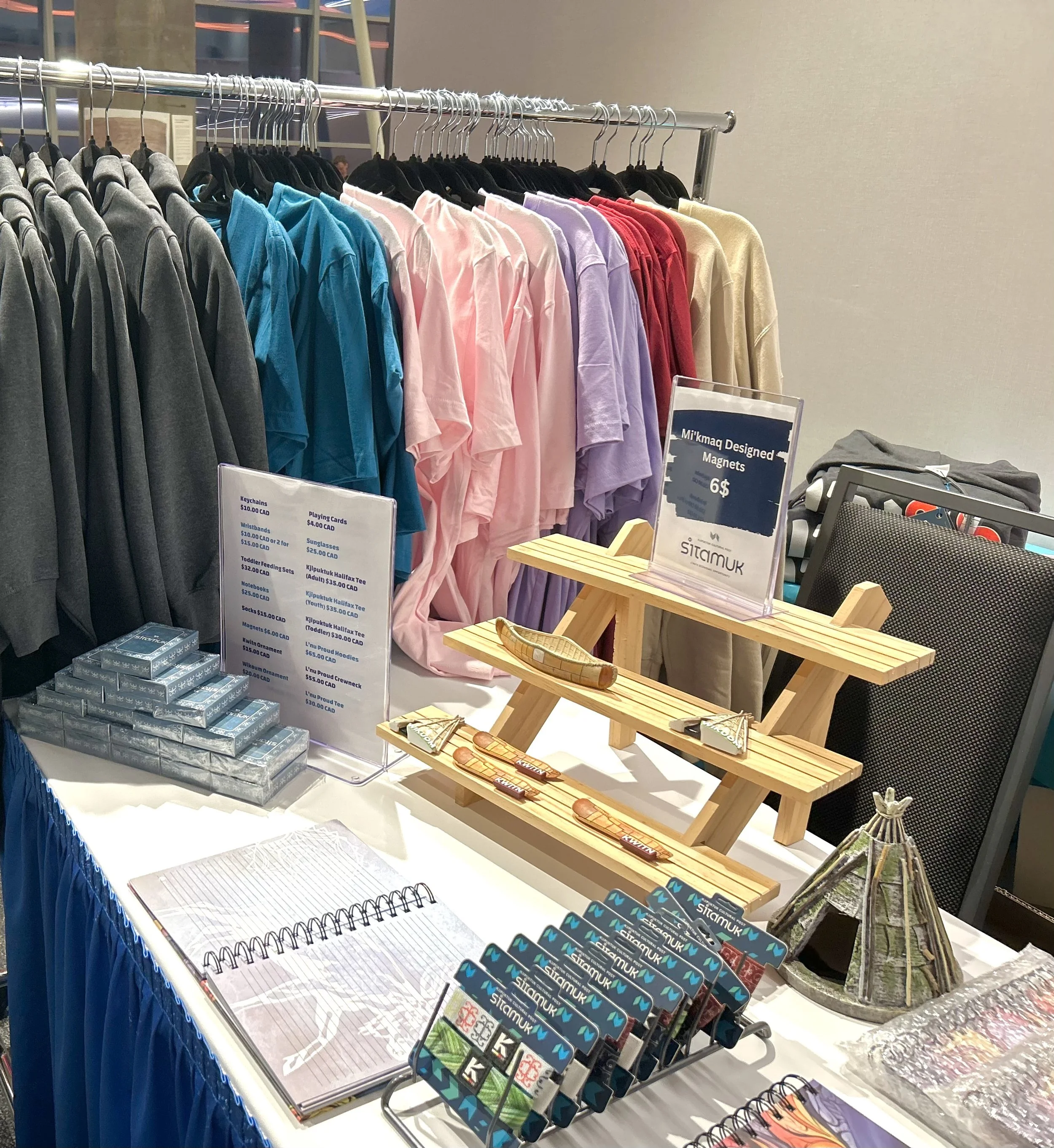 Display of colorful T-shirts on a clothing rack, with merchandise such as keychains, magnets, and notebooks on a nearby table at a market or shop.