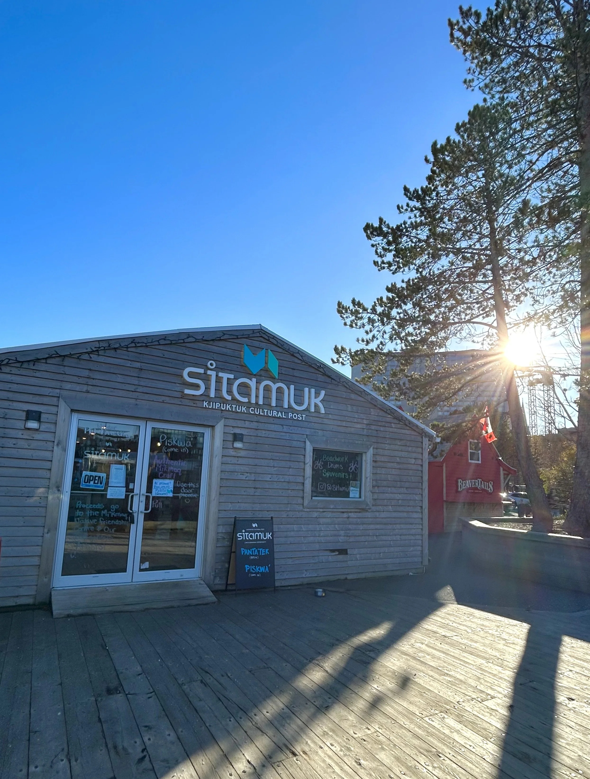 A small wooden building with a sign that reads 'Sitamuk' and 'Kjipuktuk Cultural Post,' with a tree and the sun setting in the background.