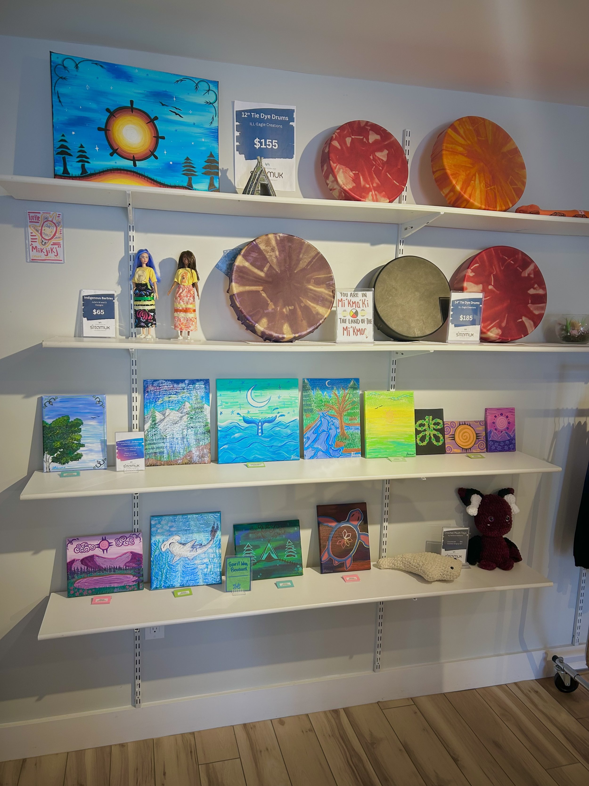 Arts and crafts display shelf with colorful circular drums on the top shelf, small paintings and dolls on the middle shelf, and additional artworks and plush toys on the bottom shelf.