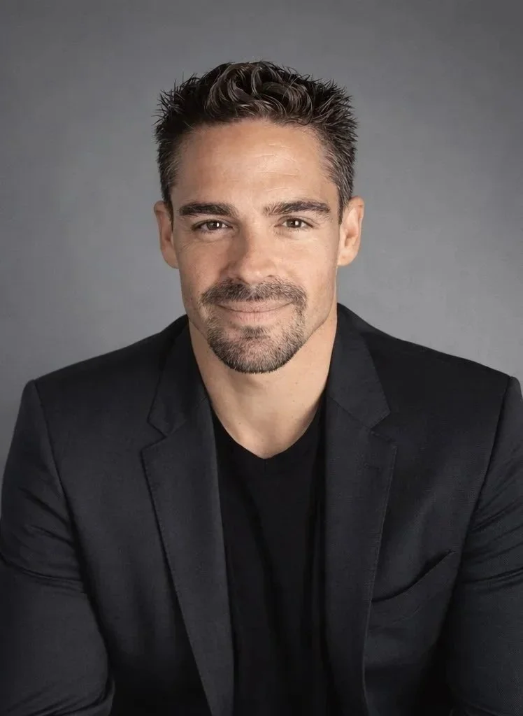 Portrait of a man with short dark hair, slight beard, wearing a black blazer and black shirt, smiling against a gray background.