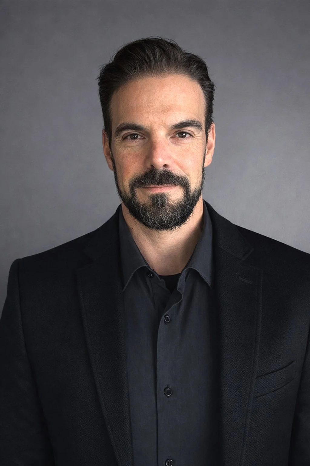A man with dark hair, a beard, and mustache, wearing a dark blazer and shirt, against a neutral gray background.