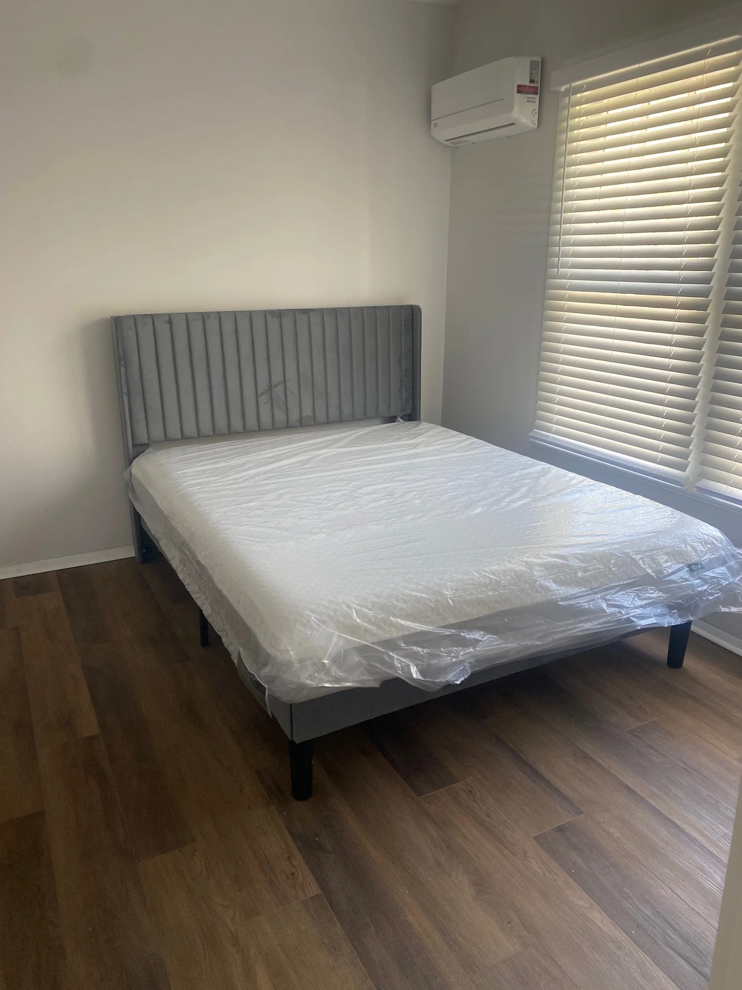 A bedroom with a gray upholstered bed frame, a mattress covered with plastic, hardwood floors, a window with blinds, and an air conditioning unit on the wall.