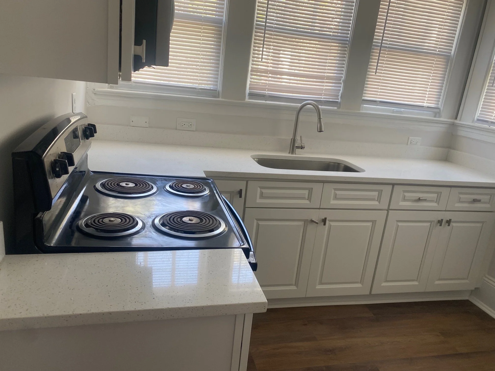 A kitchen with white cabinets, a white countertop, a stainless steel stove with four burners, a stainless steel sink, a modern faucet, and three windows with blinds.