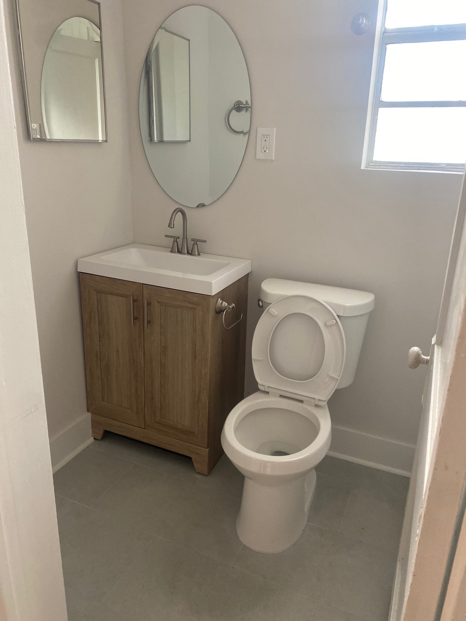 A small bathroom with a white toilet and a wooden cabinet with a white sink. There are two mirrors above the sink, a window on the wall, and a light switch.