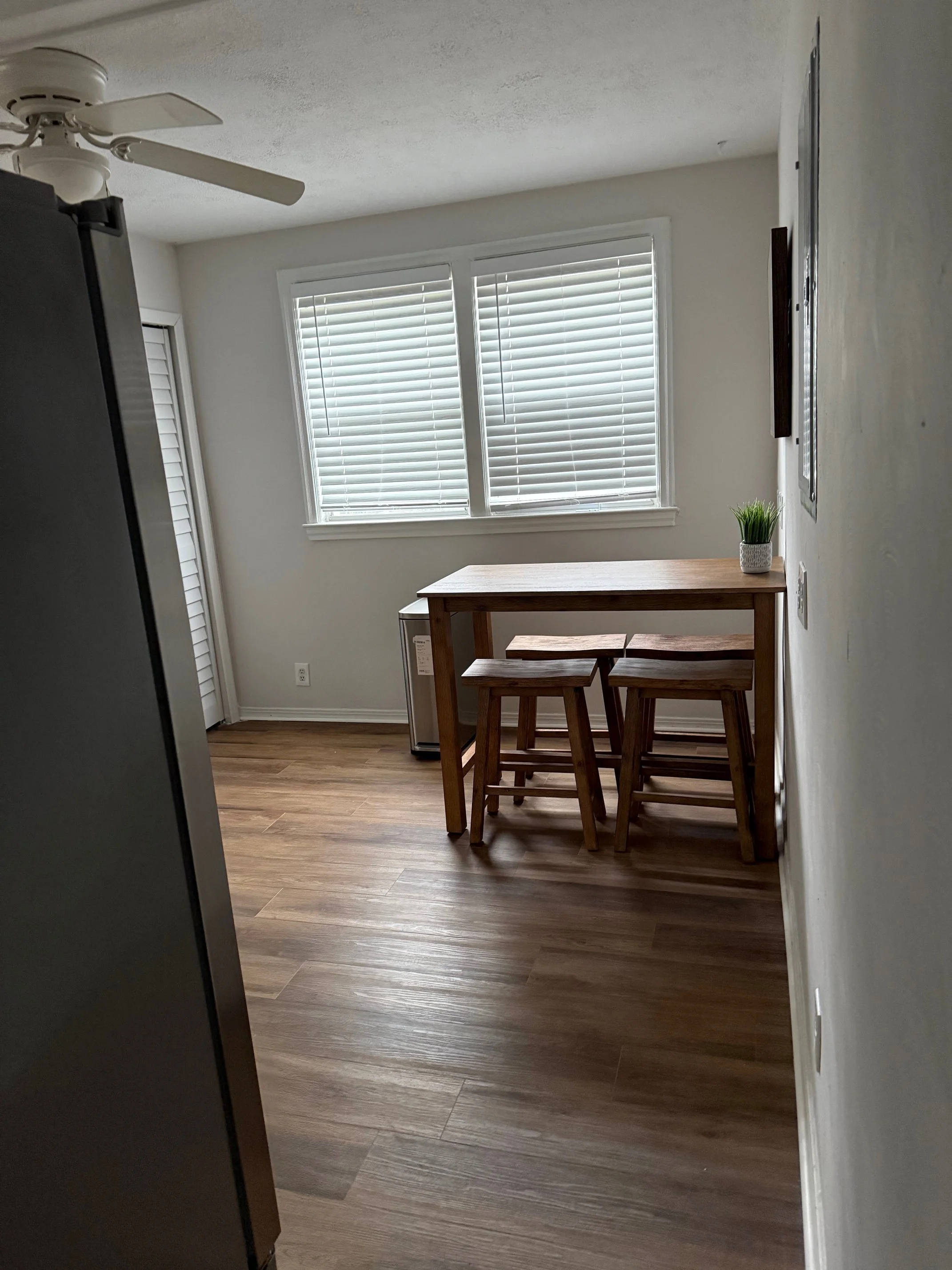 A small dining area with a wooden table and two chairs, a potted plant on the table, large windows with blinds, and a wooden floor, in a well-lit room.