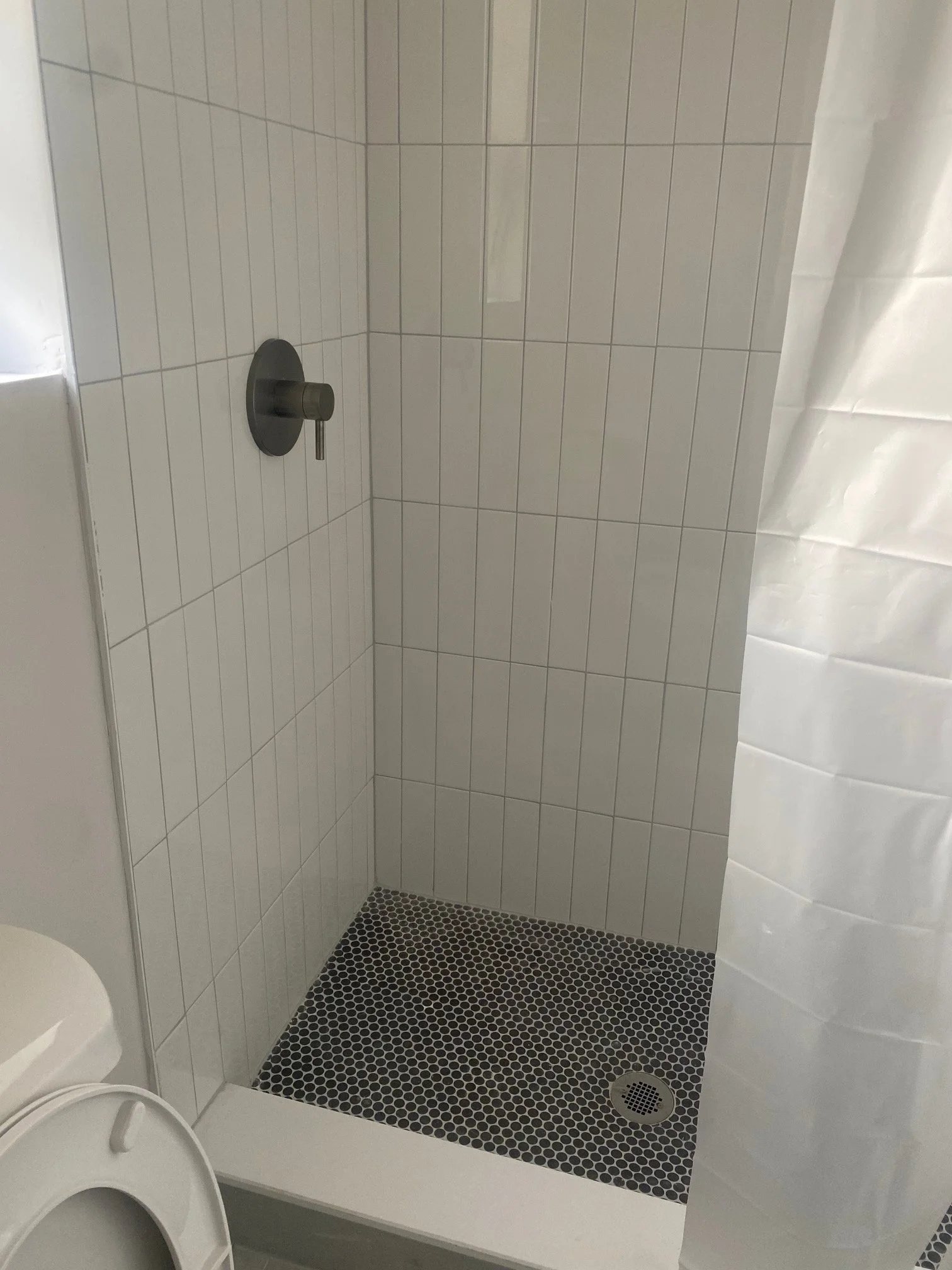 Empty shower with white tiled walls, black hexagonal tile floor, and a round drain, with a visible shower handle and a partially visible white curtain.
