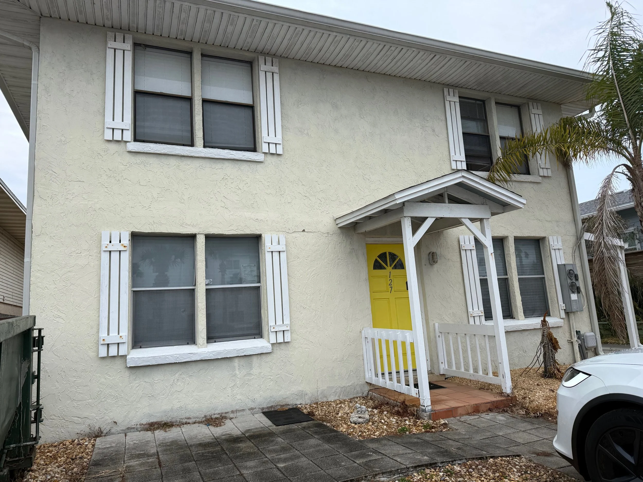 A two-story house with pale yellow stucco exterior, white window shutters, and a yellow front door with house number 127. The house has a small front porch with a white railing and a gabled roof, and there is a white car parked to the right.