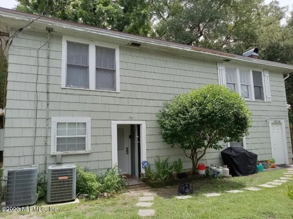Front view of a two-story house with light-colored siding, four windows, a front door, and a small tree in the yard. There are two air conditioning units and garden supplies near the house.