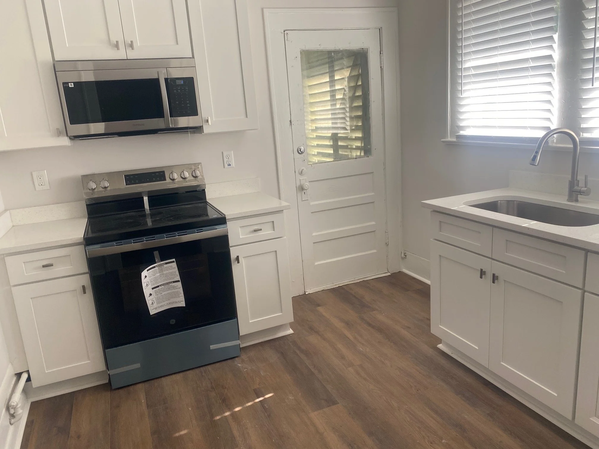 A kitchen with white cabinets, a black and stainless steel stove, a microwave, a white countertop, a sink with a chrome faucet, a window with blinds, and a wooden floor.
