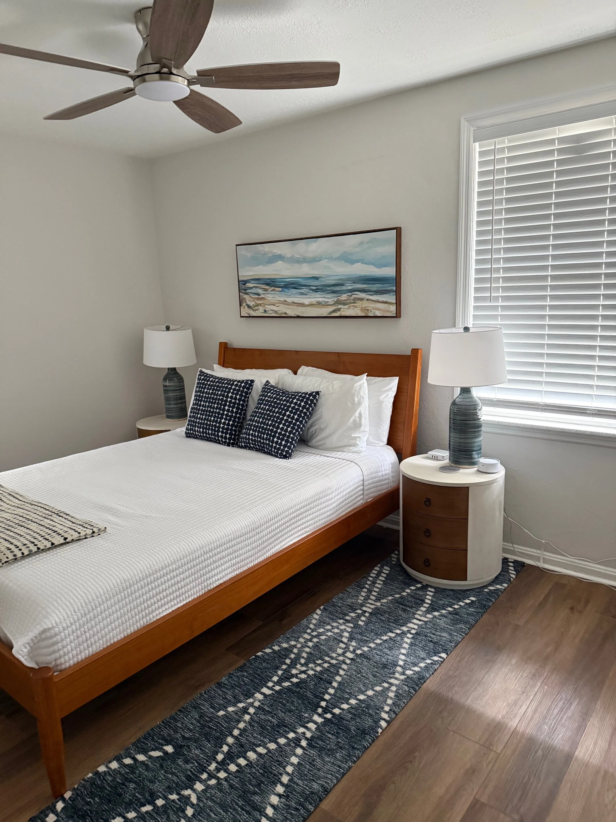 A bedroom with a wooden bed, white bedding, and black and white pillows, two nightstands with lamps, a window with blinds, a painting above the bed, a ceiling fan, and a patterned rug on wood flooring.